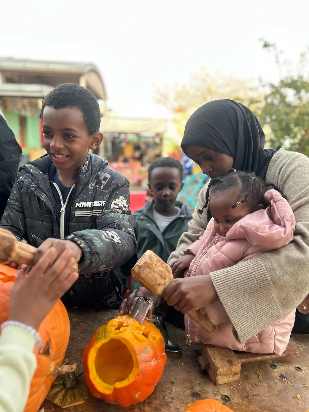 Children carving pumpkins at an outdoor activity session with buildings and autumn vegetables visible.