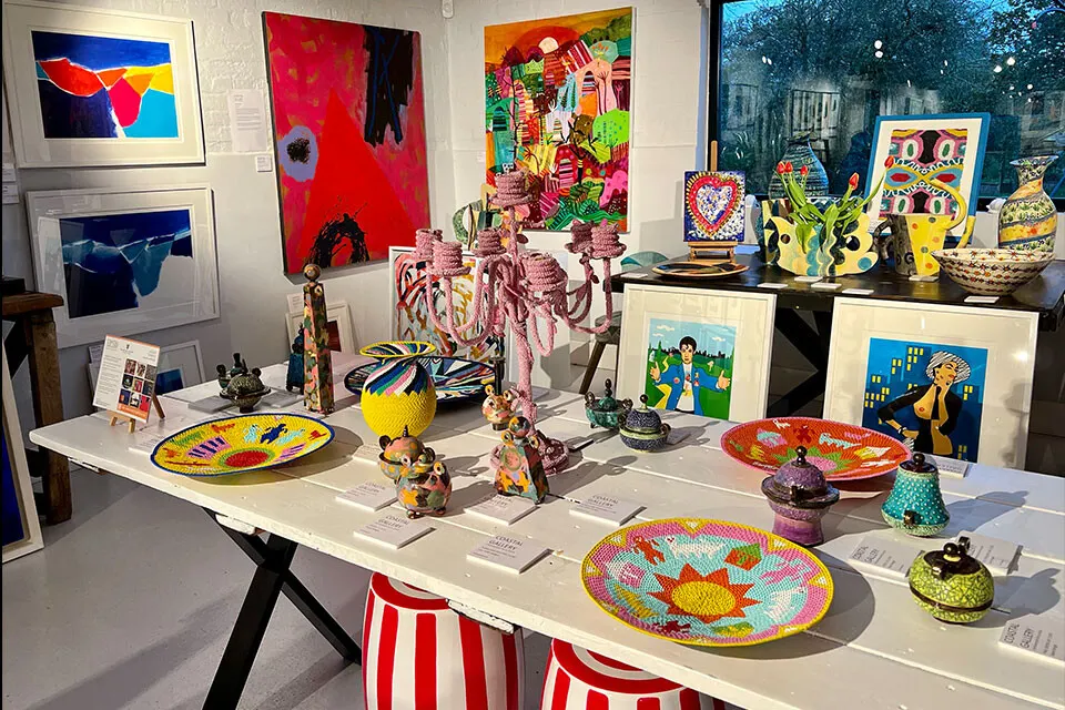 An art gallery display table showcases colourful ceramic plates, painted artworks, and decorative objects with vibrant patterns and African-inspired designs.