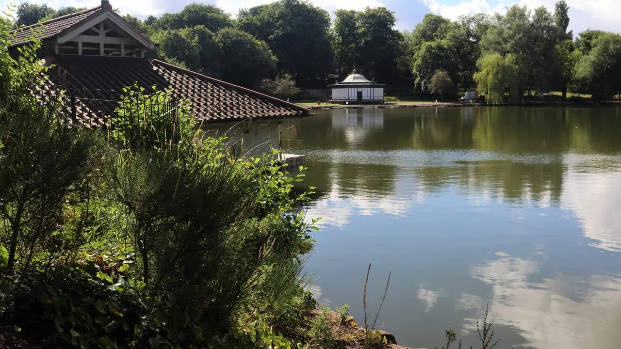 A calm lake with reedy banks, historic boathouse on the left and pavilion on the right, surrounded by mature trees.