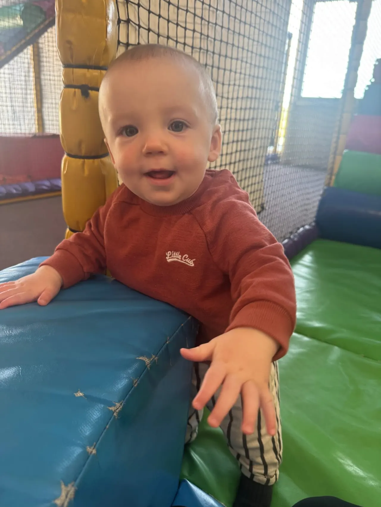 Toddler in a red jumper playing on coloured padded mats inside an indoor soft play area.