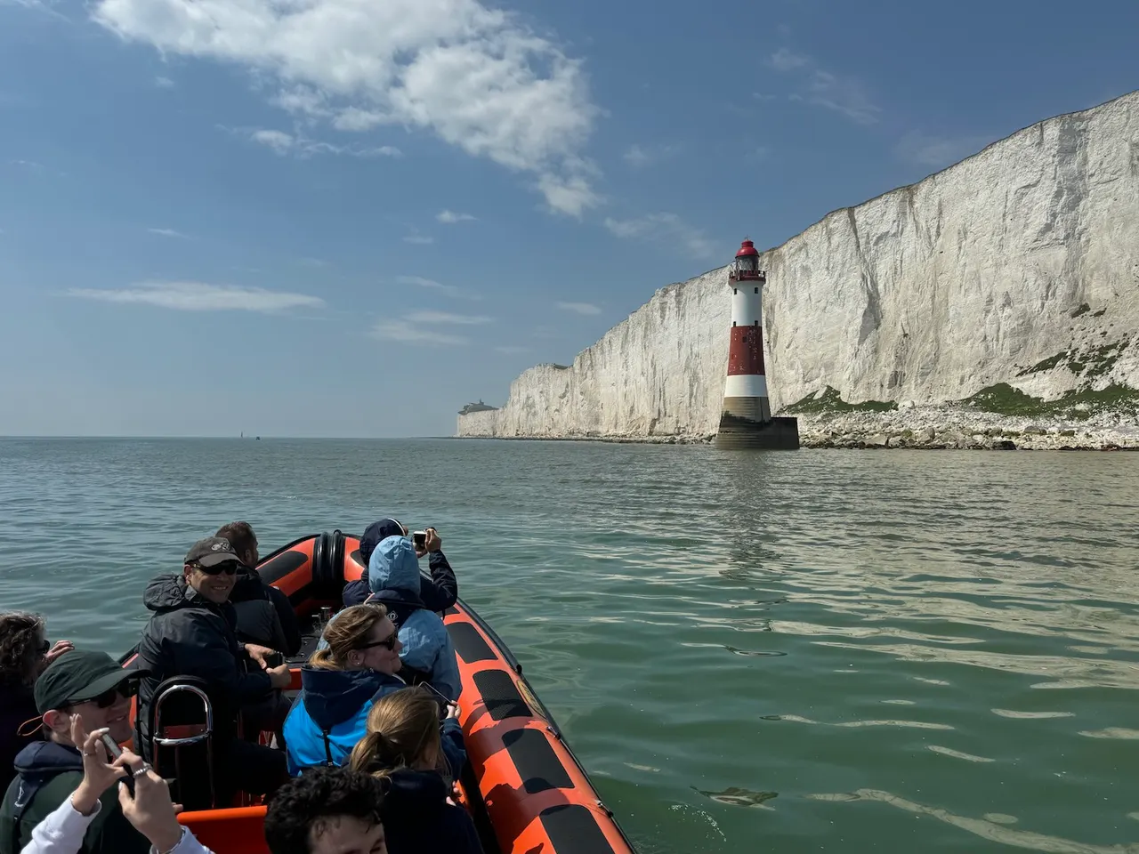 Passengers in a rigid inflatable boat view white chalk cliffs and a red-and-white striped lighthouse in calm sea.
