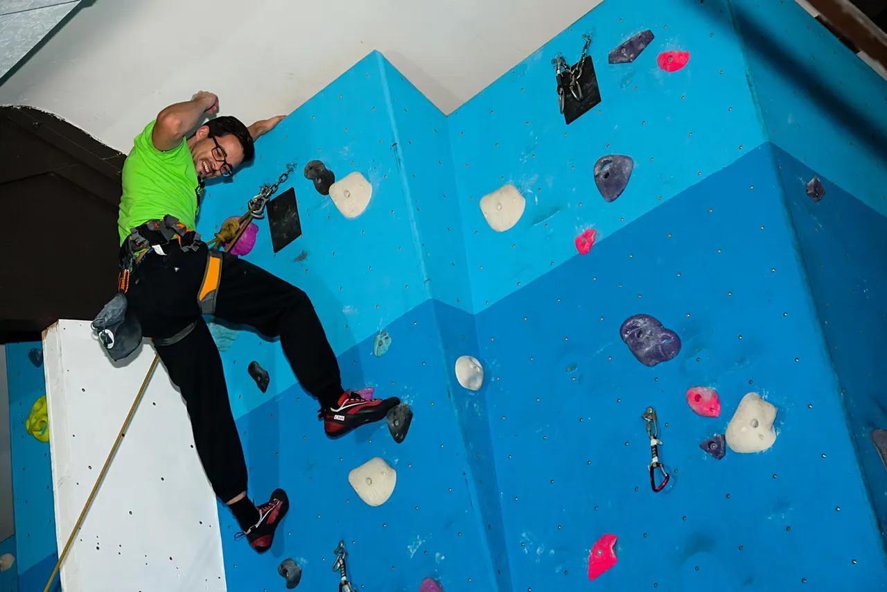 Person in bright green shirt and safety harness climbing blue indoor wall with coloured holds.