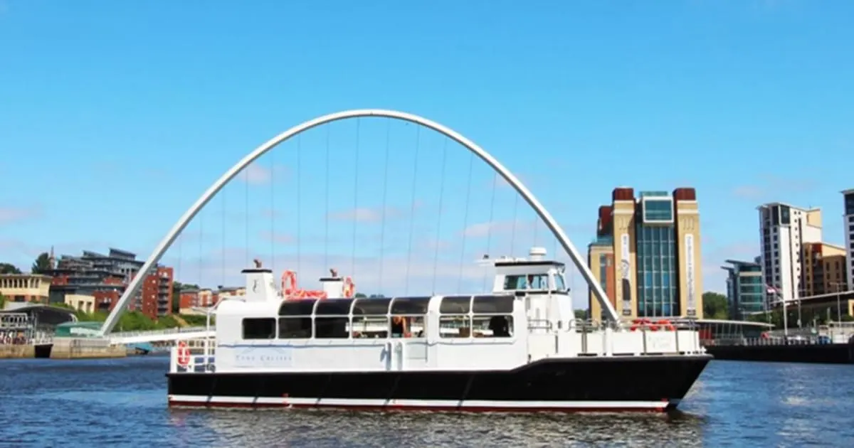White passenger riverboat on the River Tyne with the Millennium Bridge arching overhead under blue skies.