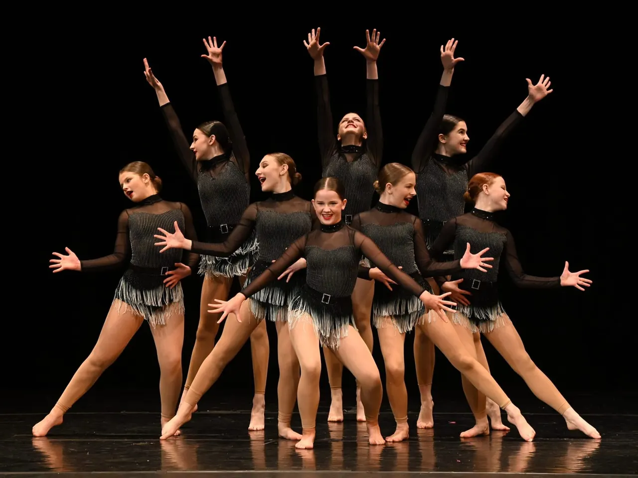 Group of young dancers performing choreographed routine on stage in black fringed costumes with arms raised.