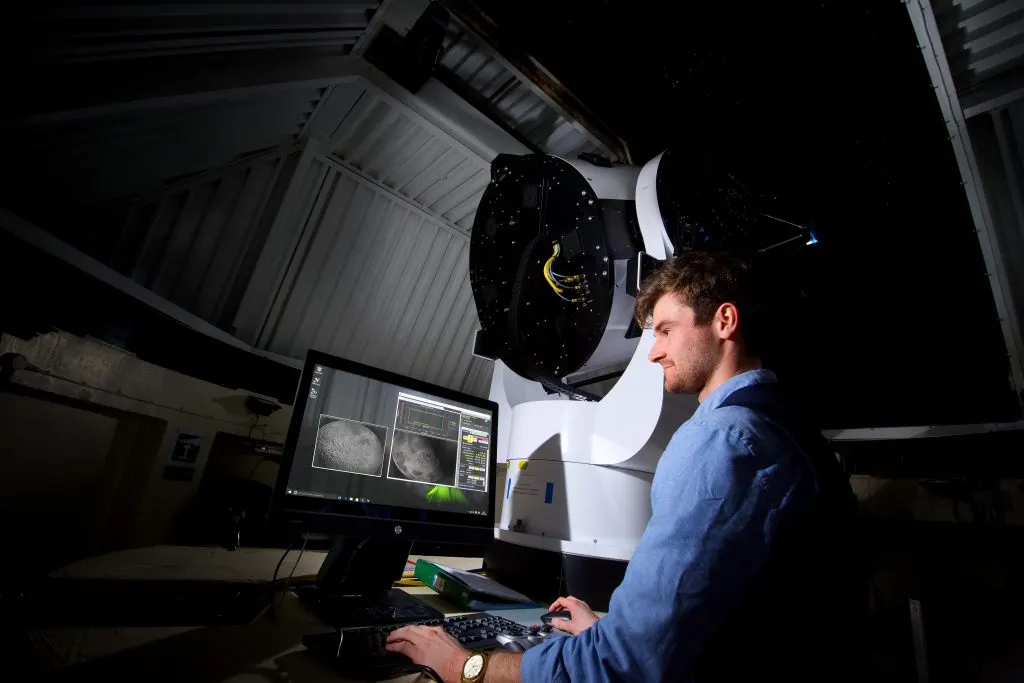 Man operating a telescope at night, viewing celestial images displayed on a computer monitor.