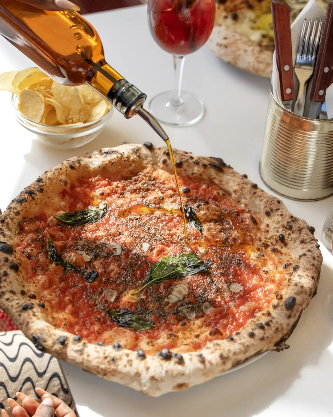 Wine being poured over a wood-fired pizza topped with tomato sauce and basil, surrounded by drinks, cutlery and snacks.