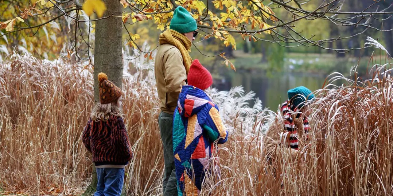 Two adults and a child walking through autumn reeds by a woodland pond, with colourful winter hats and clothing.
