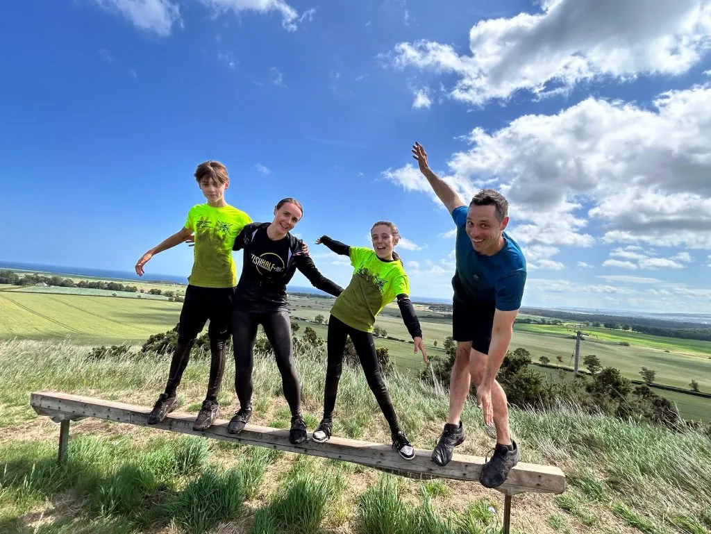 Four people in athletic wear balancing on a wooden log atop a hillside with expansive countryside views under blue sky with white clouds.