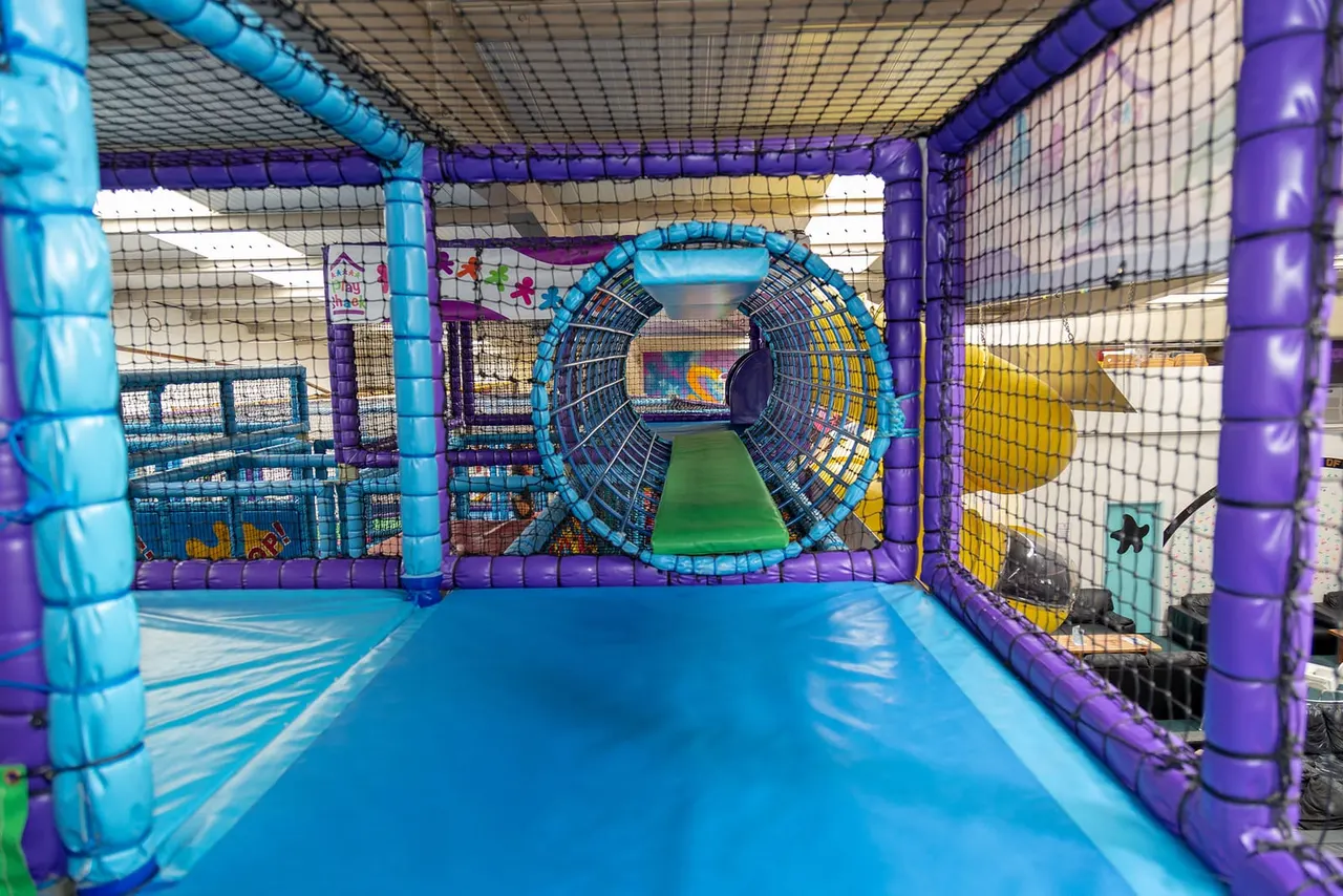 Indoor soft play structure with blue inflatable tunnels, purple pillars, and a central circular tube feature.