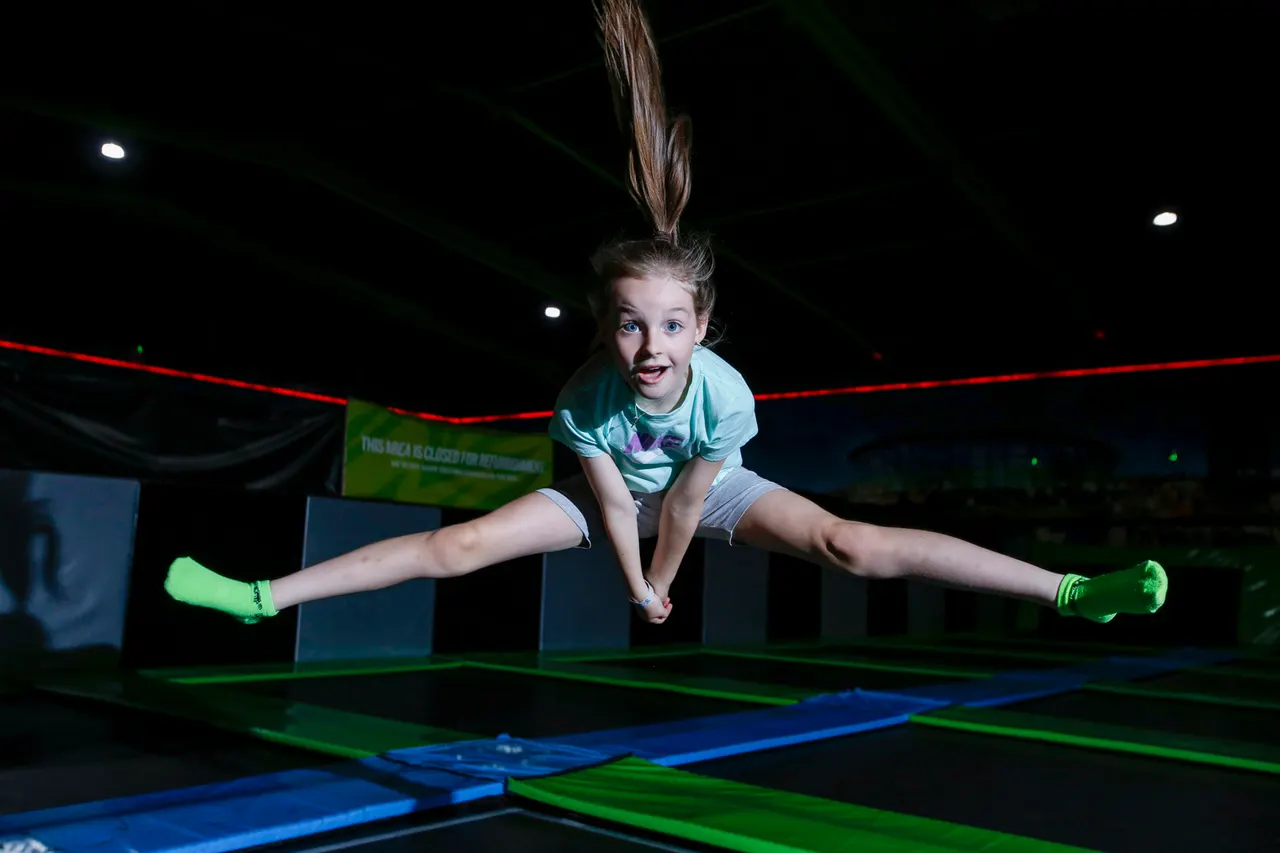 Child bouncing on a trampoline with legs stretched wide, wearing bright green socks in an indoor trampoline park.