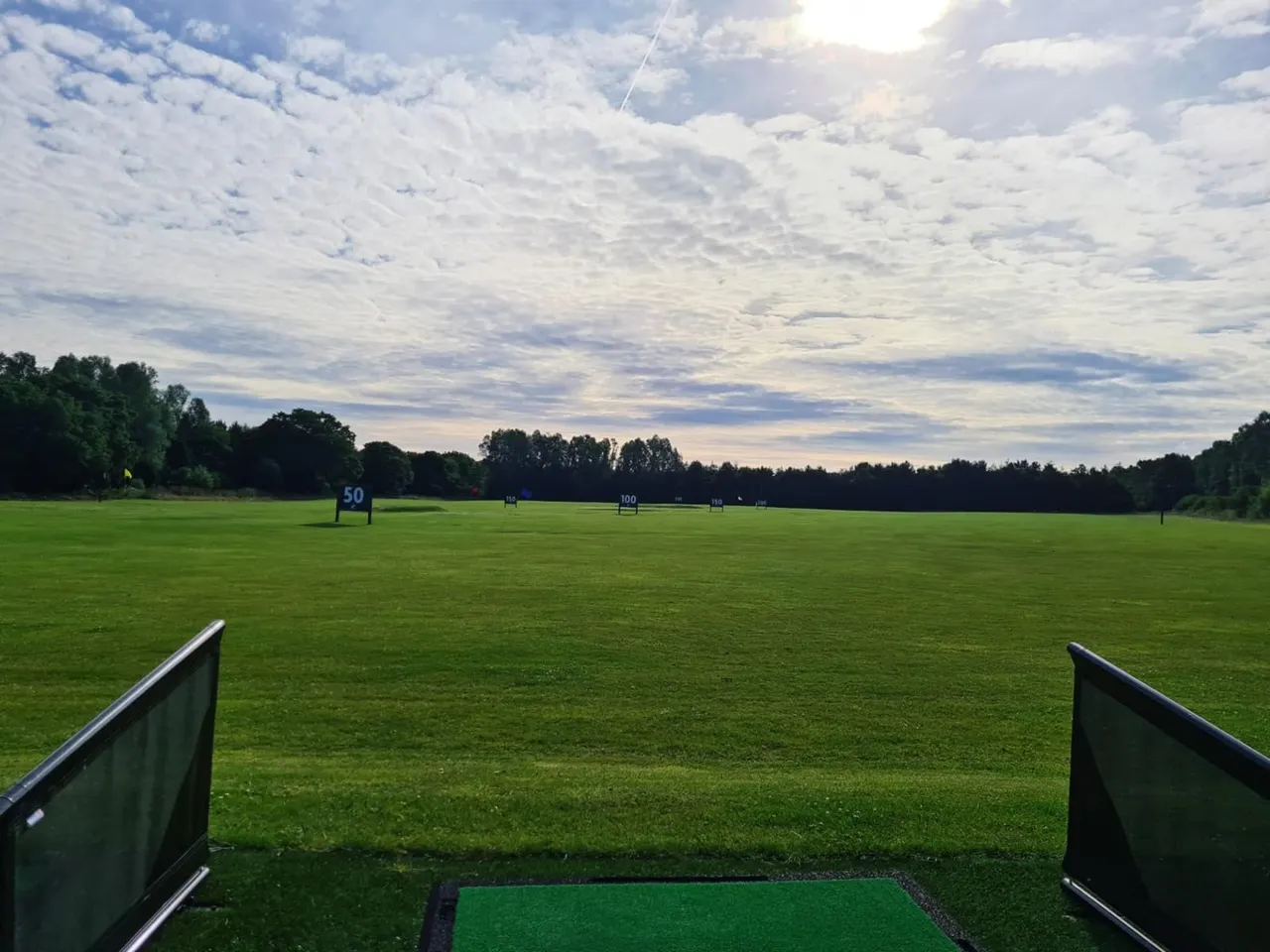 Open grass field with distance markers and wooded boundaries under a cloudy sky, viewed from a golf tee.