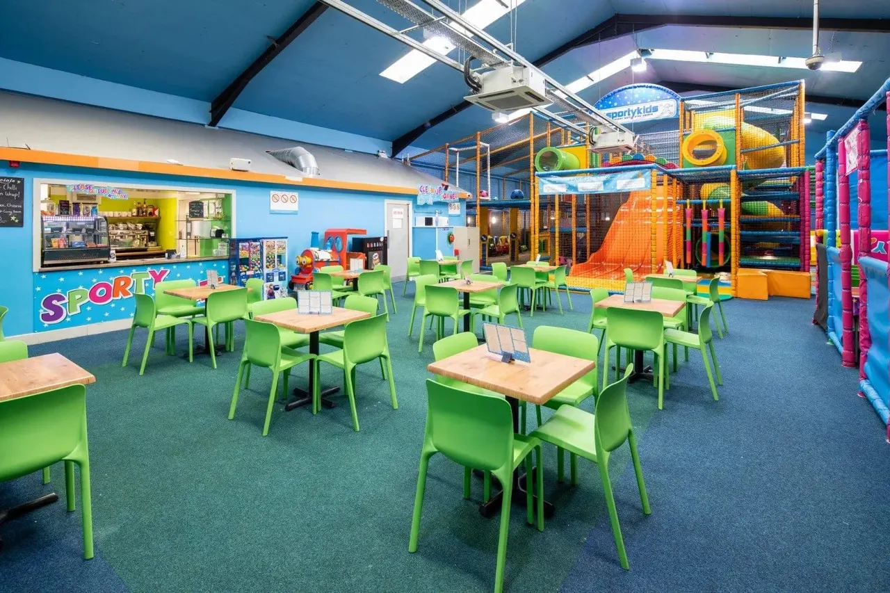 Indoor children's play venue with dining area featuring bright green chairs and wooden tables, colourful climbing structures and slides in background.