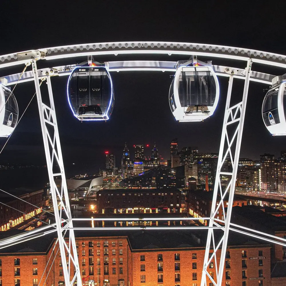 Illuminated observation wheel pods suspended above Liverpool's lit-up cityscape and waterfront at night.