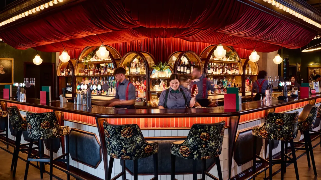 Bar staff serve behind an ornate wooden counter beneath a dramatic red velvet canopy in a vintage-style cocktail bar.
