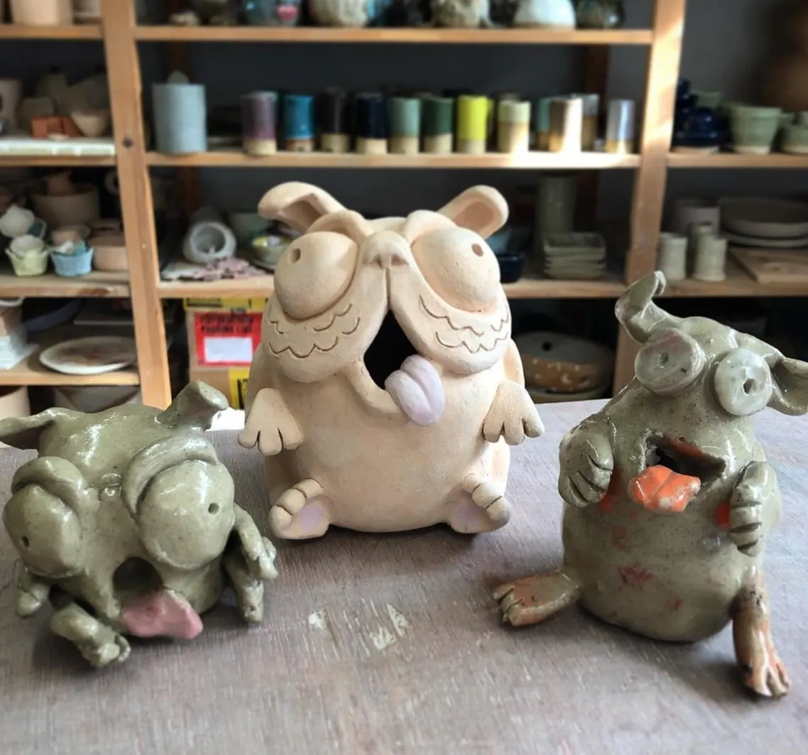 Handmade ceramic animal figurines displayed on a worktable in a pottery studio, with shelves of tools and materials behind.