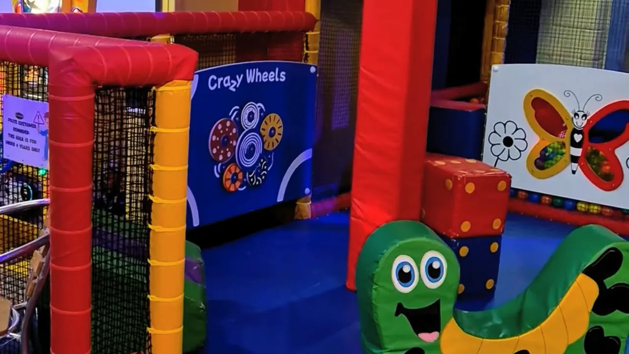 Colourful indoor soft play area with inflatable structures, the "Crazy Wheels" ride, and cartoon character decorations.