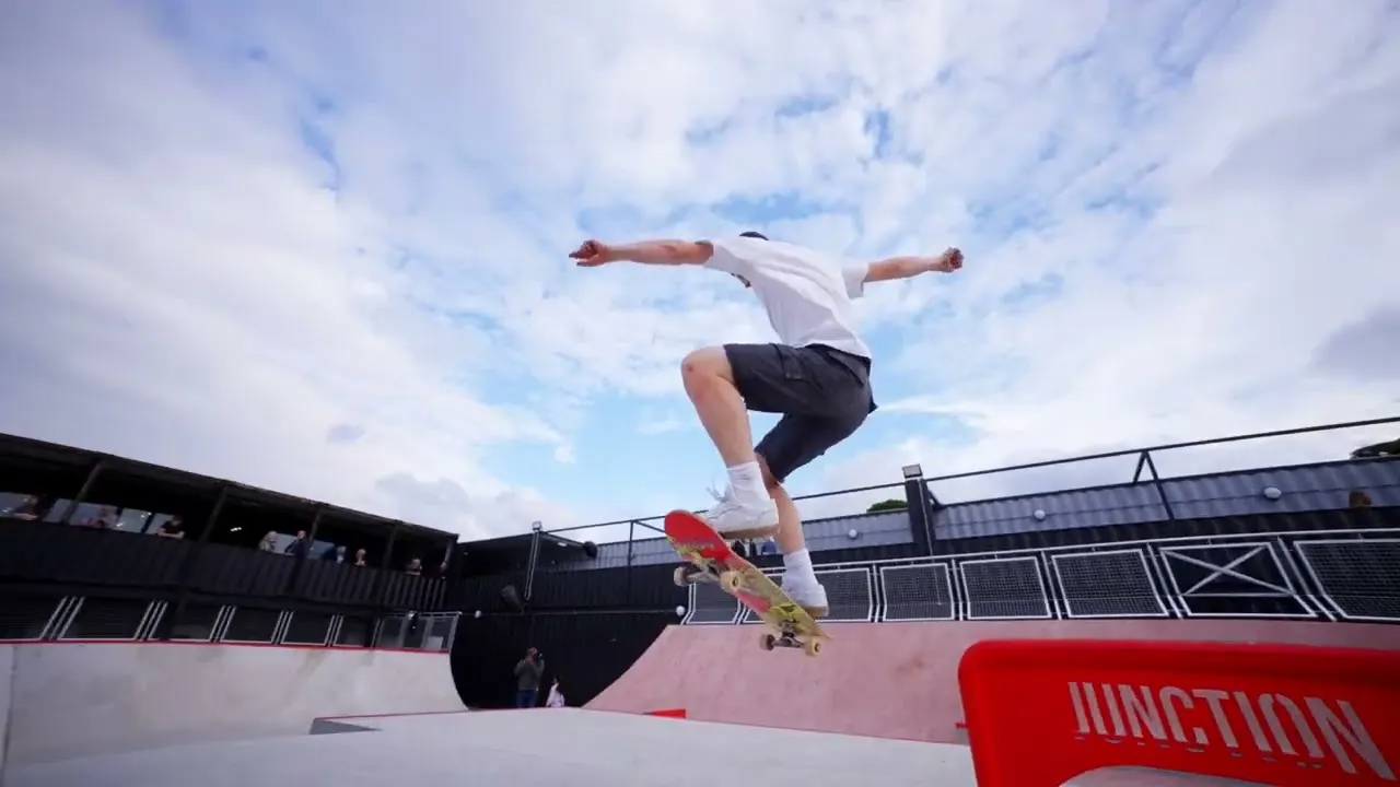 A skateboarder performs a trick mid-air at an outdoor skate park with spectators watching from the stands.