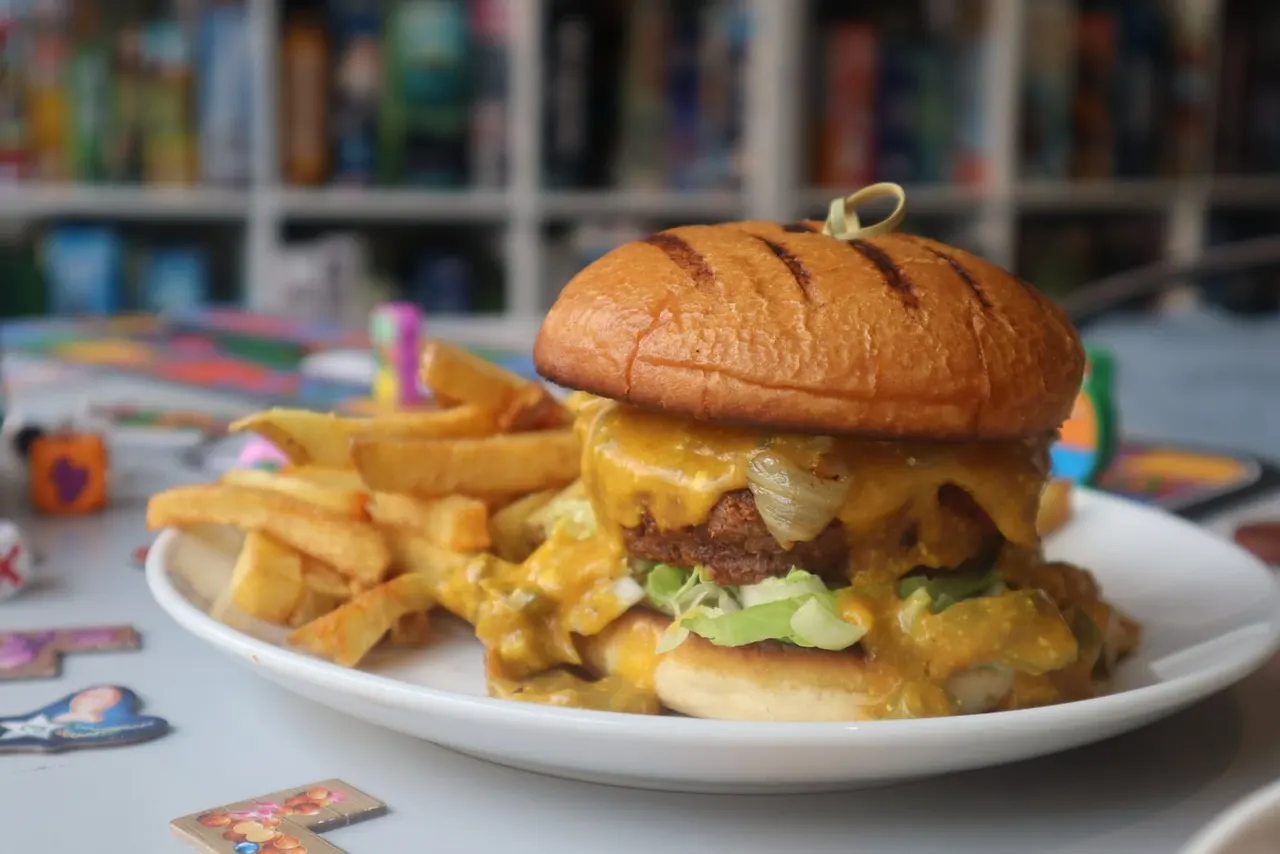 Cheeseburger with melted cheese and lettuce served with chips on a white plate, with board game pieces visible nearby.