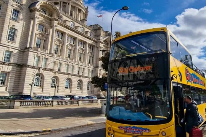 Yellow open-top tour bus with passengers on a city street beside a grand stone government building.