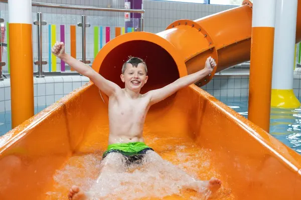 Young child emerging from orange water slide with arms raised in an indoor swimming pool.
