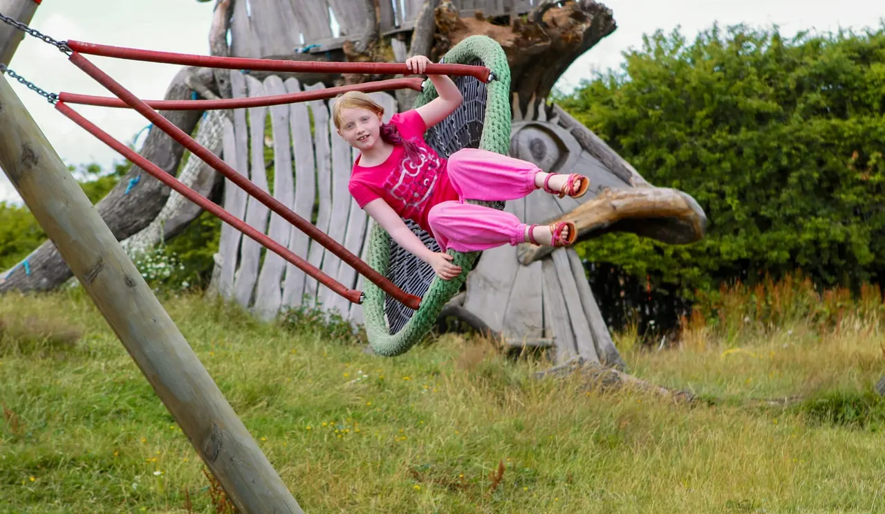 Young child in pink outfit playing on wooden climbing frame with red bars in outdoor playground.