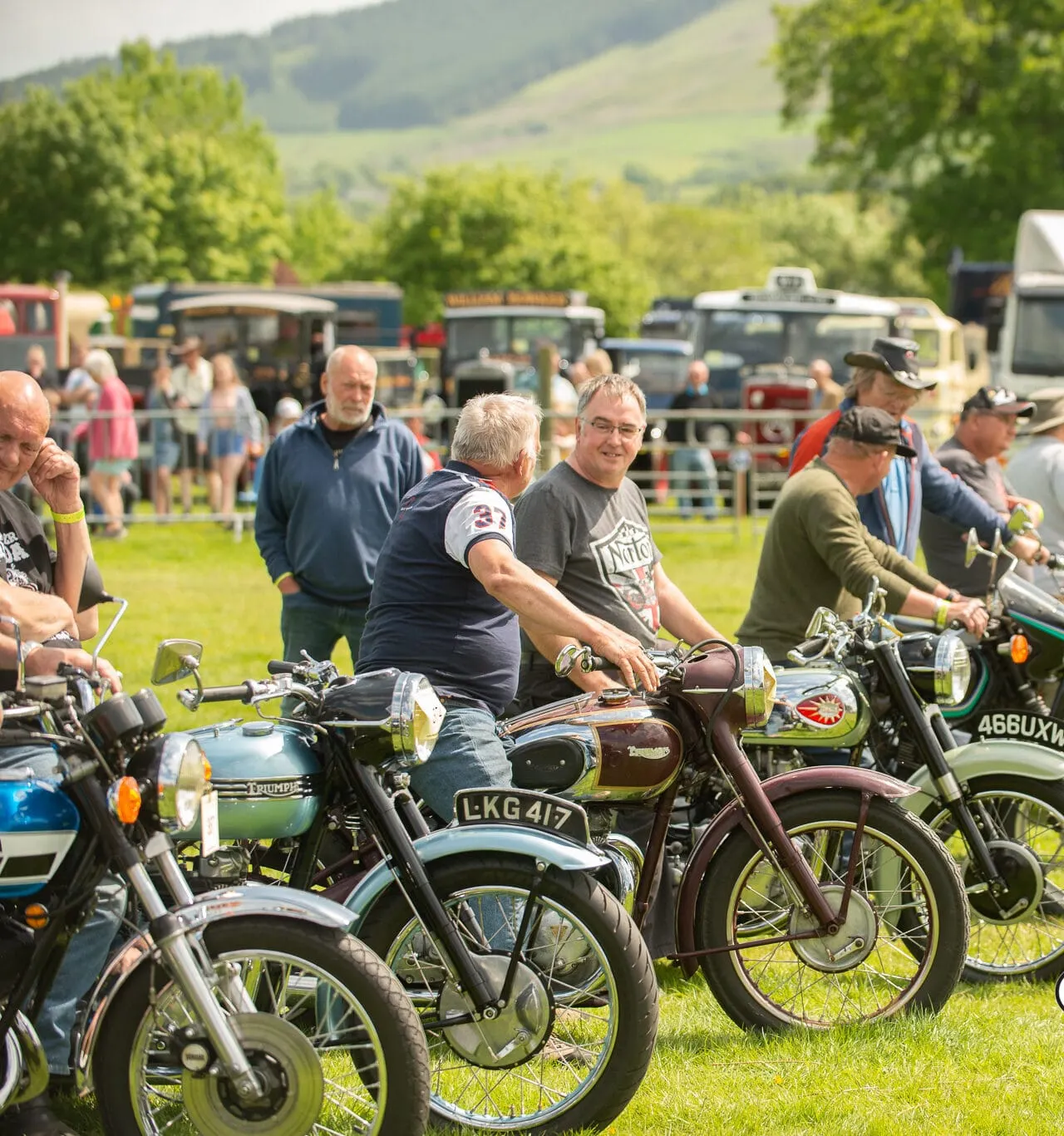 Vintage motorcycles on display at an outdoor rally with visitors and rolling countryside hills in the background.