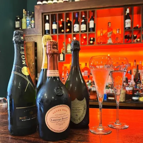 Three champagne bottles and two filled flute glasses arranged on a red bar counter with illuminated orange shelving behind.