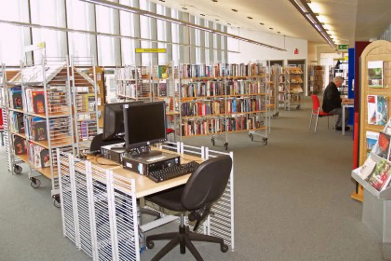 Library interior with computer workstation in foreground, book-lined shelves, and visitors browsing collections.