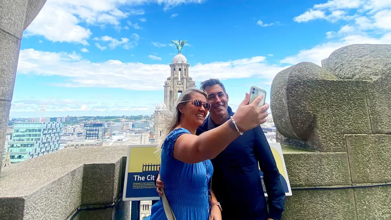 Two visitors taking a selfie on an observation deck with a cityscape and historic tower in the background.