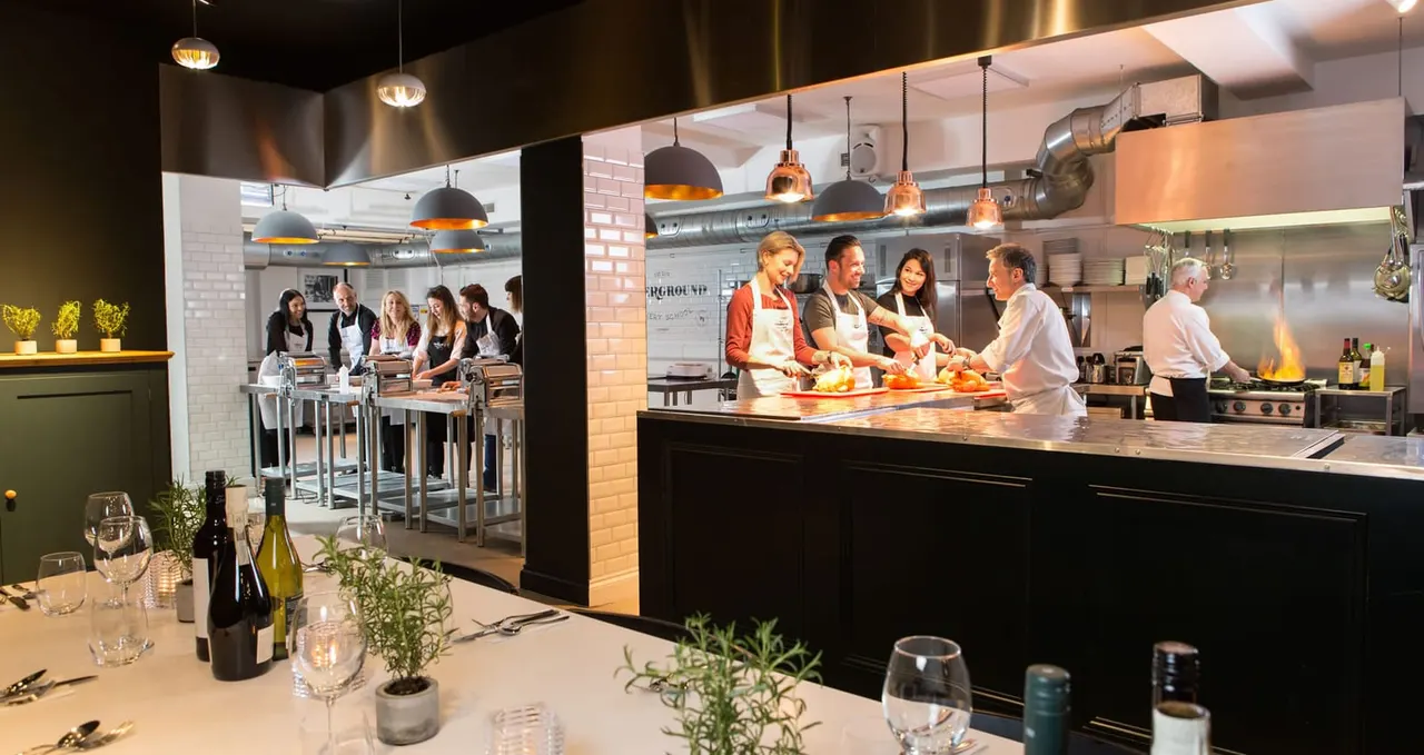 Staff and participants engaged in a cooking class at an open kitchen counter with diners seated at tables in the foreground.