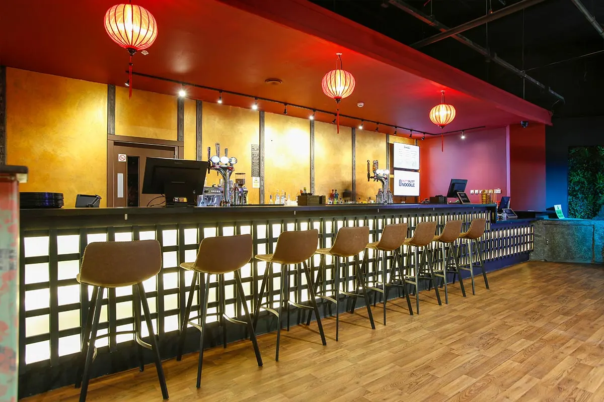 Bar counter with stools in a colourfully lit venue with hanging lanterns and wooden flooring.