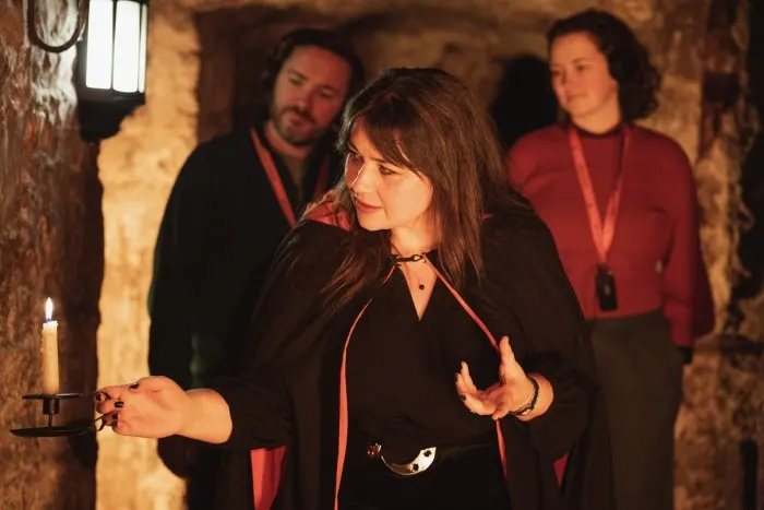 A guide in black jacket gestures whilst speaking to visitors in a stone-walled underground chamber.