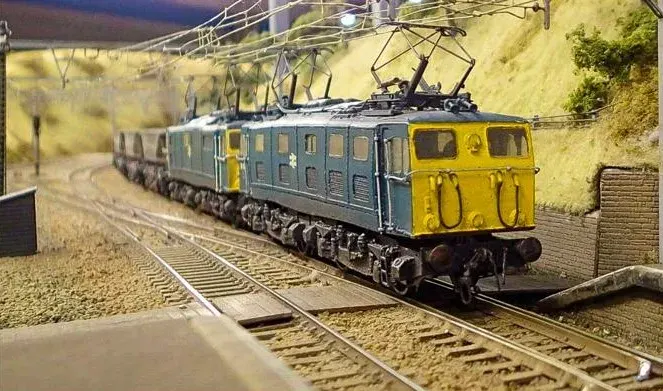 Model railway layout featuring a yellow and green electric locomotive pulling passenger carriages along miniature track.