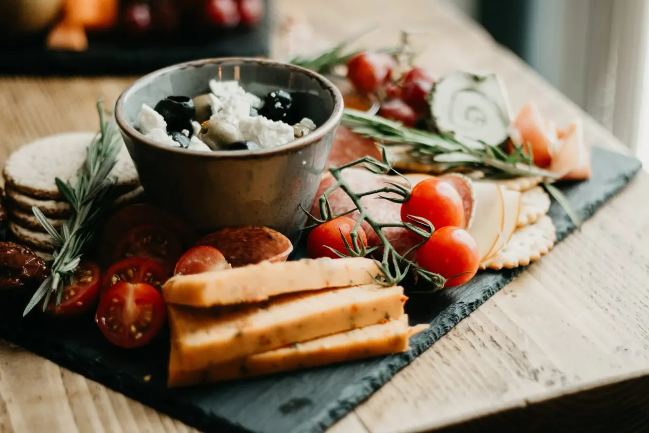 Mediterranean charcuterie board with cheese, cured meats, tomatoes, olives, rosemary and crackers on slate.