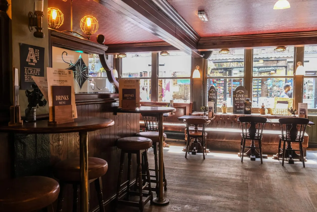 Interior of a traditional pub with wooden bar, bar stools, tables and chairs, warm lighting, and large windows overlooking a street.