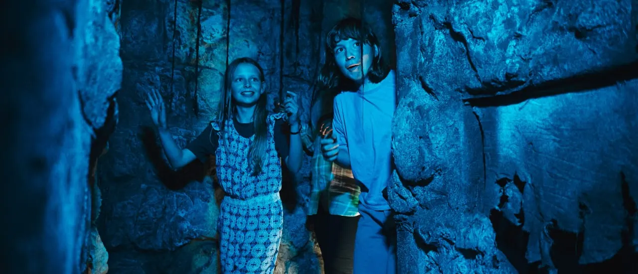 Two children explore a dark cave or mine tunnel lit with blue lighting.