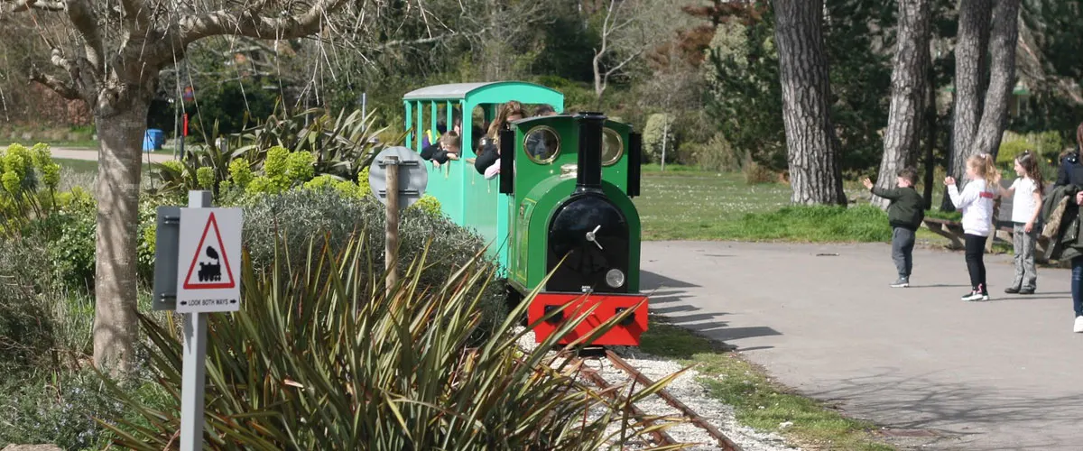 A miniature green and red steam train with passengers moves along a track through a tree-lined park, with visitors walking nearby.