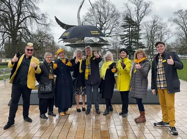 A group of adults wearing yellow and black clothing give thumbs up whilst standing together under a large umbrella sculpture on a paved outdoor area, with bare trees in the background.
