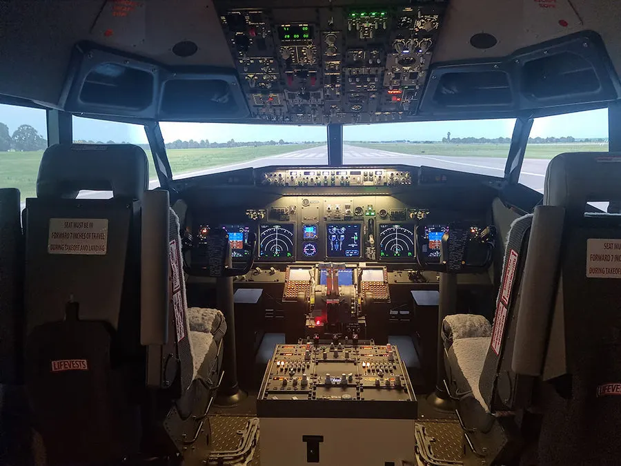 Cockpit interior of a flight simulator with control panels, two pilot seats, and a runway visible through the front windows.