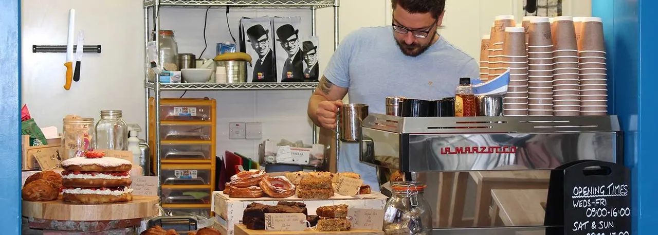 Man operating espresso machine behind counter with cakes and pastries displayed in front.