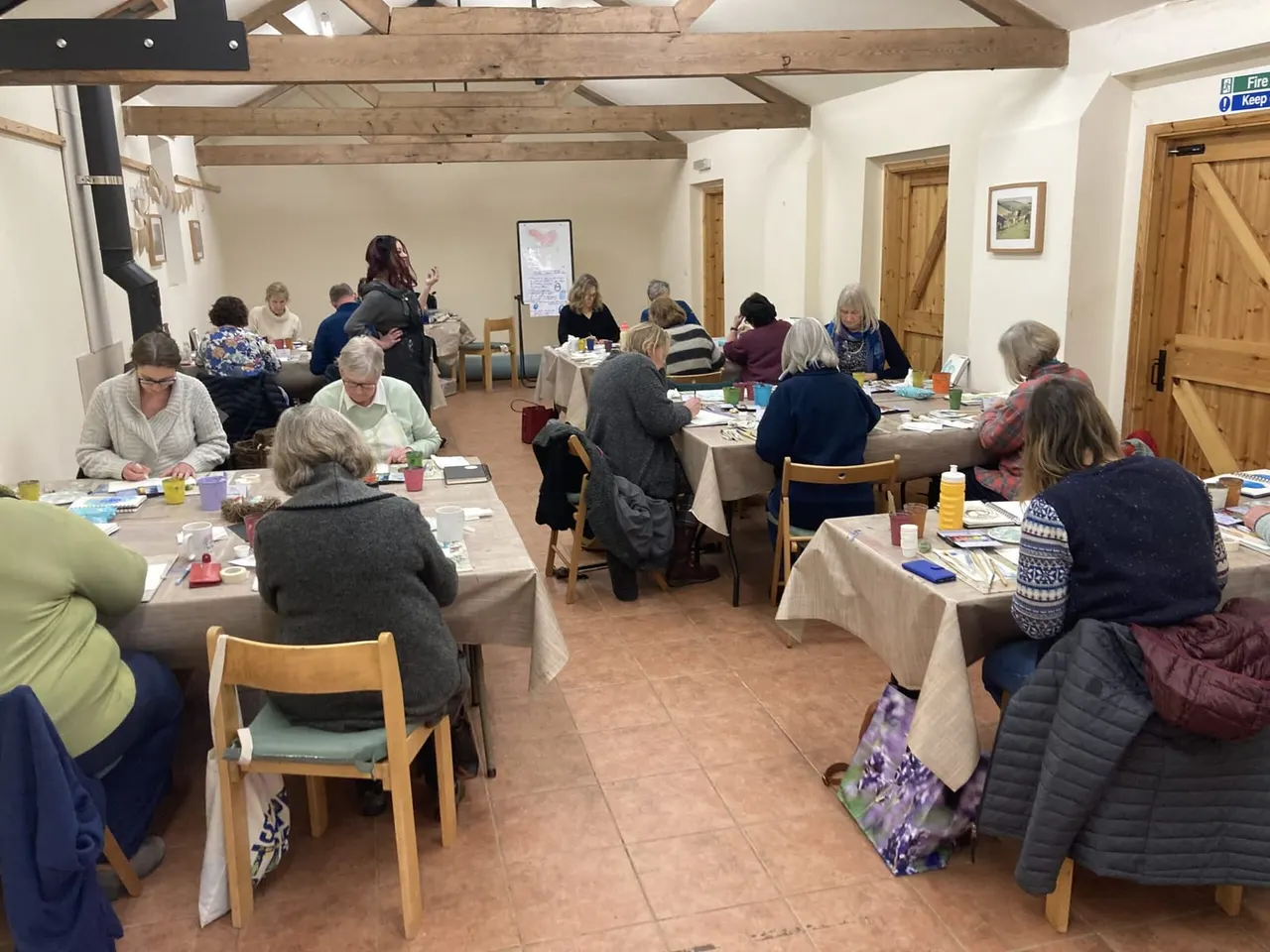 A group of adults seated at tables during an art class, with craft materials and refreshments, in a room with exposed wooden beams.