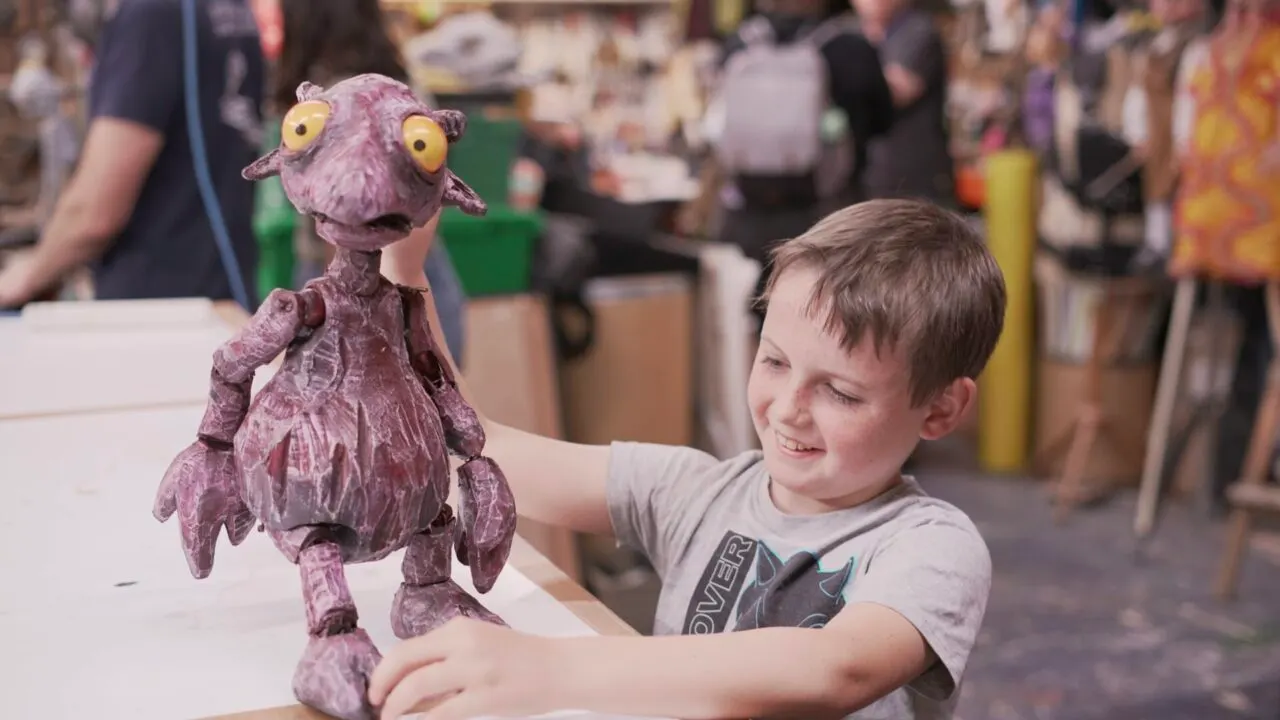 A young child smiles while holding a purple puppet with yellow eyes at a busy indoor venue.