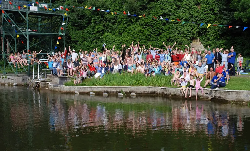 A large crowd of visitors gathered along the waterside at Henleaze Lake, many with arms raised, watching activities by the water with colourful bunting overhead.