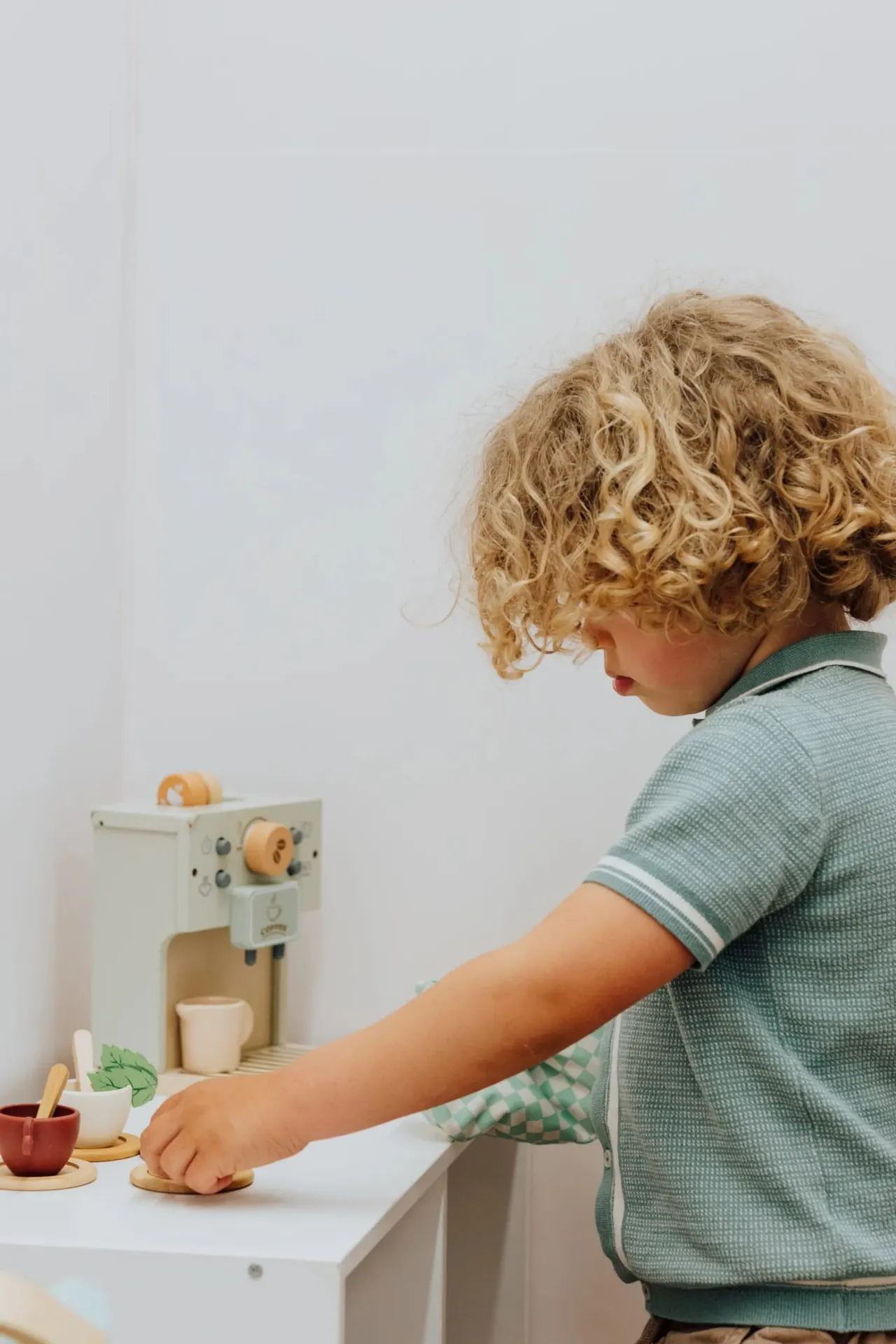 Child with curly blonde hair playing with a toy wooden coffee machine at a white table.