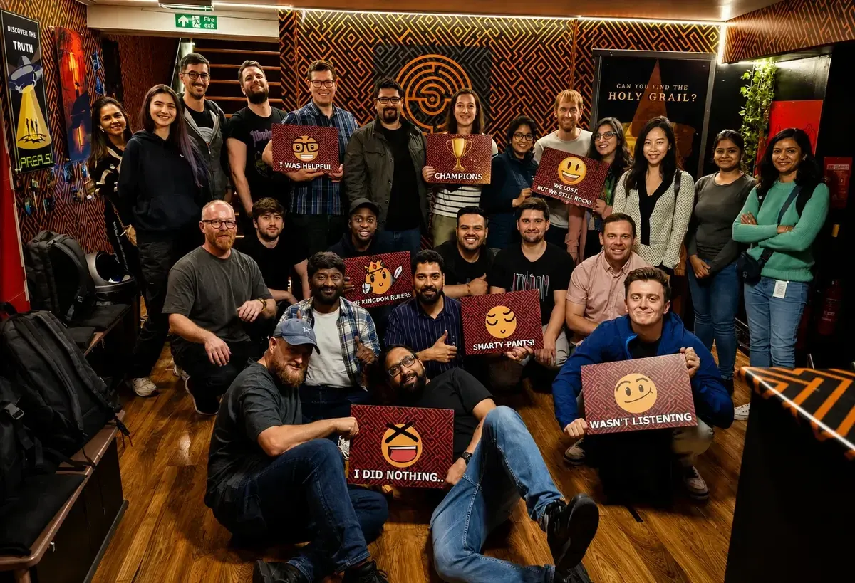 Large group of people posing together indoors holding game boards with humorous messages in an escape room venue.