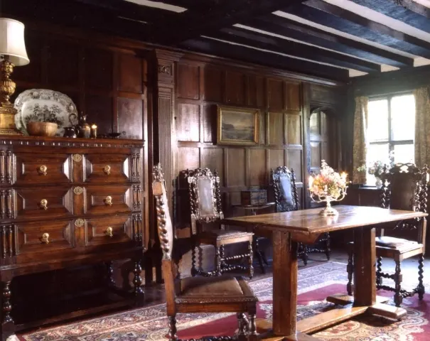 Historic interior room with wooden furniture, panelled walls, a large table, ornate chest of drawers, and period decorations.