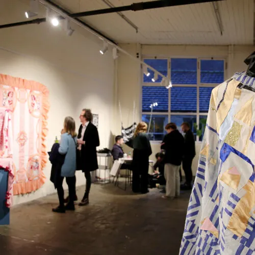 Visitors viewing textile artwork displayed on walls and hanging fabrics in an industrial gallery space.