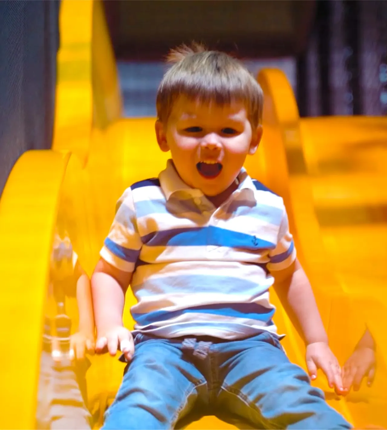 A young child in a striped shirt sits on a bright yellow inflatable slide, smiling with mouth open.
