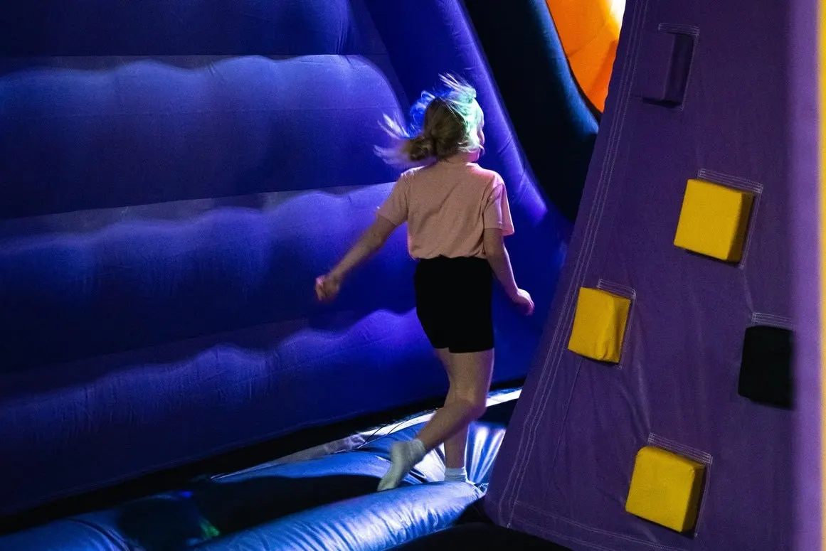Child bouncing on illuminated blue inflatable structures inside a trampoline park.