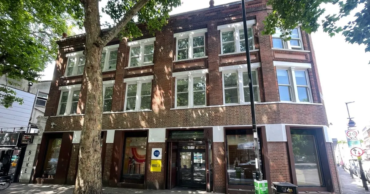 Red-brick building with ground-floor shopfront and residential floors above, shaded by a large tree.